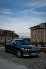 A luxury black car is parked on a cobblestone driveway in front of elegant, beige buildings with red-tiled roofs. The sky is partially cloudy, casting a serene atmosphere over the scene.