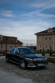 A luxury black car is parked on a cobblestone driveway in front of elegant, beige buildings with red-tiled roofs. The sky is partially cloudy, casting a serene atmosphere over the scene.