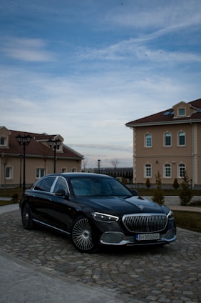 A luxury black car is parked on a cobblestone driveway in front of elegant, beige buildings with red-tiled roofs. The sky is partially cloudy, casting a serene atmosphere over the scene.