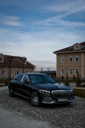 A luxury black car is parked on a cobblestone driveway in front of elegant, beige buildings with red-tiled roofs. The sky is partially cloudy, casting a serene atmosphere over the scene.