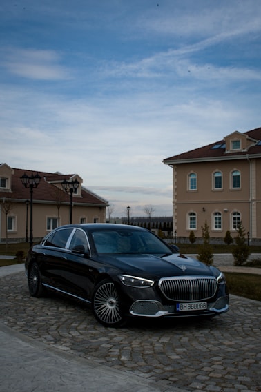 A luxury black car is parked on a cobblestone driveway in front of elegant, beige buildings with red-tiled roofs. The sky is partially cloudy, casting a serene atmosphere over the scene.