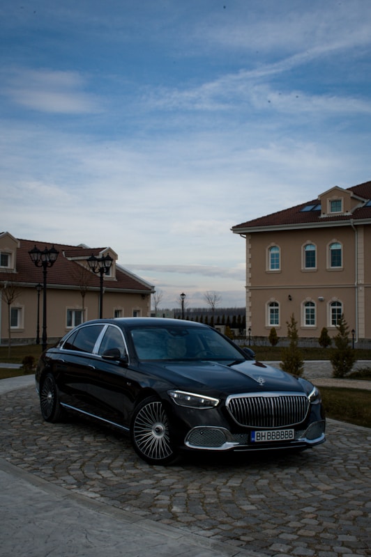 A luxury black car is parked on a cobblestone driveway in front of elegant, beige buildings with red-tiled roofs. The sky is partially cloudy, casting a serene atmosphere over the scene.