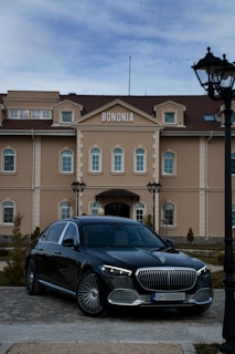 A sleek black luxury car is parked in front of an elegant building with the sign 'BONONIA' on top. The building features beige walls, arched windows, and multiple levels with a symmetrical design. Well-manicured shrubs and a cobblestone path are also visible in the foreground, and there is a street lamp on the right.