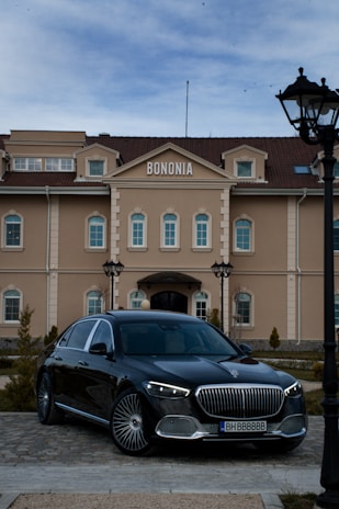 A sleek black luxury car is parked in front of an elegant building with the sign 'BONONIA' on top. The building features beige walls, arched windows, and multiple levels with a symmetrical design. Well-manicured shrubs and a cobblestone path are also visible in the foreground, and there is a street lamp on the right.