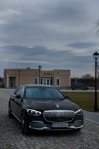 A luxury car with striking headlights is parked on a cobblestone driveway. In the background, there is a beige building labeled 'SHOP' with lamp posts lining the path. The sky is overcast, creating a moody atmosphere.