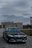 A luxury car with striking headlights is parked on a cobblestone driveway. In the background, there is a beige building labeled 'SHOP' with lamp posts lining the path. The sky is overcast, creating a moody atmosphere.