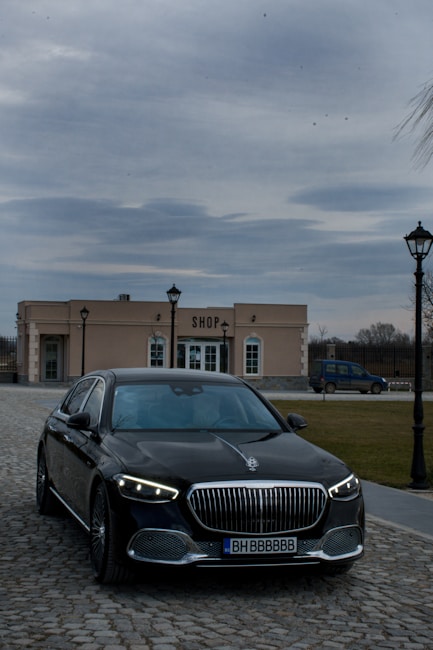 A luxury car with striking headlights is parked on a cobblestone driveway. In the background, there is a beige building labeled 'SHOP' with lamp posts lining the path. The sky is overcast, creating a moody atmosphere.