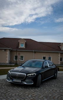 A luxurious black car is parked on a cobblestone path in front of a large house with a brown tiled roof. The sky above is partly cloudy, adding depth to the scene.