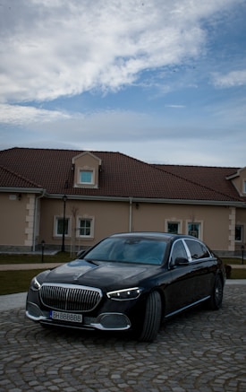 A luxurious black car is parked on a cobblestone path in front of a large house with a brown tiled roof. The sky above is partly cloudy, adding depth to the scene.