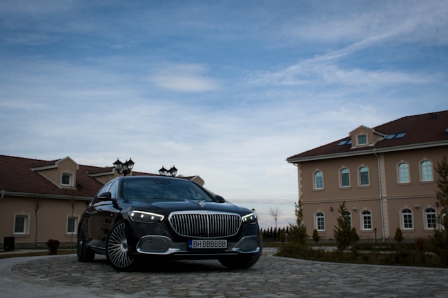 A luxury car is parked in front of large, elegant buildings with beige facades. The sky is mostly clear with some clouds visible. The car is sleek and polished, parked on a cobblestone driveway leading towards the buildings.