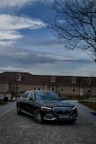 A luxury car with a sleek design is parked on a cobblestone road in front of a large house. The sky is partly cloudy with a bare tree on the right side, adding a serene yet sophisticated atmosphere.
