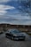 A luxury car with a sleek design is parked on a cobblestone road in front of a large house. The sky is partly cloudy with a bare tree on the right side, adding a serene yet sophisticated atmosphere.