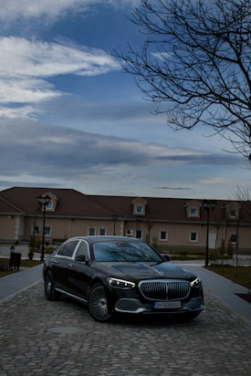 A luxury car with a sleek design is parked on a cobblestone road in front of a large house. The sky is partly cloudy with a bare tree on the right side, adding a serene yet sophisticated atmosphere.