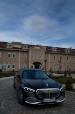 A luxury car is parked in front of an elegant building named 'Bononia'. The building displays classic architectural design with multiple windows and a symmetrical facade. The sky above is partially cloudy.