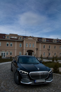 A luxury car is parked in front of an elegant building named 'Bononia'. The building displays classic architectural design with multiple windows and a symmetrical facade. The sky above is partially cloudy.