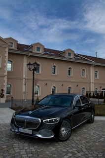 A luxurious black car is parked on a cobblestone driveway in front of an elegant beige building with multiple windows. The sky is clear with a few clouds, and a decorative streetlamp is visible beside the car.