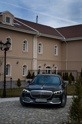 A luxury black car is parked in front of a beige-colored building with multiple windows and a brown tiled roof. The foreground shows a cobblestone driveway lined with small conical shrubs, and street lamps stand to the left. The sky above is slightly overcast.