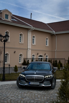 A luxury black car is parked in front of a beige-colored building with multiple windows and a brown tiled roof. The foreground shows a cobblestone driveway lined with small conical shrubs, and street lamps stand to the left. The sky above is slightly overcast.