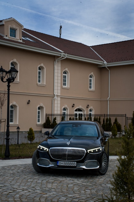 A luxury black car is parked in front of a beige-colored building with multiple windows and a brown tiled roof. The foreground shows a cobblestone driveway lined with small conical shrubs, and street lamps stand to the left. The sky above is slightly overcast.