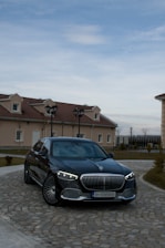 A luxury black sedan is parked on a cobblestone driveway in front of a house with beige walls and red-tiled roofs. The vehicle has distinctive LED headlights and a prominent grille. The sky is overcast with some blue patches visible.