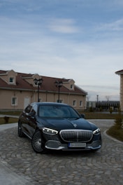 A luxury black sedan is parked on a cobblestone driveway in front of a house with beige walls and red-tiled roofs. The vehicle has distinctive LED headlights and a prominent grille. The sky is overcast with some blue patches visible.