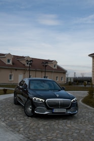 A luxury black sedan is parked on a cobblestone driveway in front of a house with beige walls and red-tiled roofs. The vehicle has distinctive LED headlights and a prominent grille. The sky is overcast with some blue patches visible.