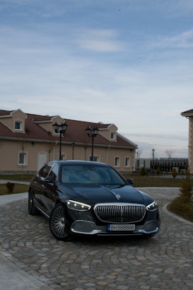 A luxury black sedan is parked on a cobblestone driveway in front of a house with beige walls and red-tiled roofs. The vehicle has distinctive LED headlights and a prominent grille. The sky is overcast with some blue patches visible.