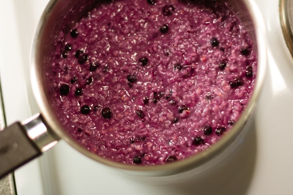 A pot filled with purple porridge featuring small dark berries throughout. The mixture has a textured appearance and is being cooked on a stove.