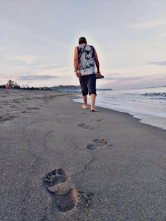 A smiling traveler walking barefoot along the shoreline with footprints in the sand.