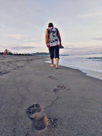 A smiling traveler walking barefoot along the shoreline with footprints in the sand.