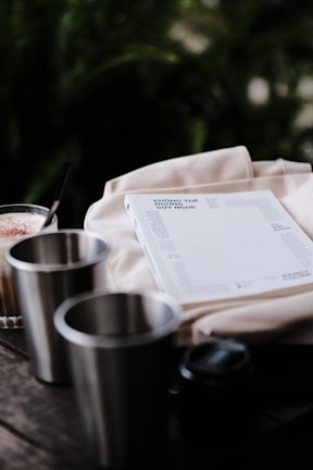 A soft, light-colored cloth serves as the backdrop for a white book with text on the cover in a foreign language. Near the book, a latte with a foamy top in a clear glass presents spoonfuls of cocoa powder or cinnamon and is ready for sipping with a black straw. Two stainless steel cups add a modern touch, while everything rests on a rustic wooden surface surrounded by a blurred green background.