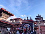 A vibrant and busy scene featuring traditional Nepalese architecture with intricately carved wooden details and pagoda-styled roofs adorned with red trims. Multiple temples are visible, and a large depiction of a deity can be seen as part of the decoration. The sky is clear and blue, contrasting with the earthy tones of the brick buildings. Crowds of people are engaging in what appears to be a cultural or religious gathering, giving a sense of activity and liveliness.