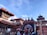 A vibrant and busy scene featuring traditional Nepalese architecture with intricately carved wooden details and pagoda-styled roofs adorned with red trims. Multiple temples are visible, and a large depiction of a deity can be seen as part of the decoration. The sky is clear and blue, contrasting with the earthy tones of the brick buildings. Crowds of people are engaging in what appears to be a cultural or religious gathering, giving a sense of activity and liveliness.