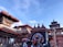 A vibrant and busy scene featuring traditional Nepalese architecture with intricately carved wooden details and pagoda-styled roofs adorned with red trims. Multiple temples are visible, and a large depiction of a deity can be seen as part of the decoration. The sky is clear and blue, contrasting with the earthy tones of the brick buildings. Crowds of people are engaging in what appears to be a cultural or religious gathering, giving a sense of activity and liveliness.