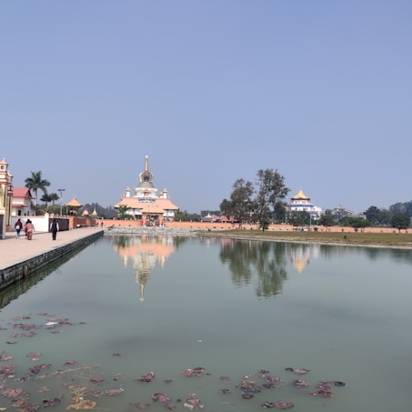 A serene temple complex with a still water body reflecting the structures and sky. The temple features ornate architecture with a tall spire and traditional design elements. Trees line the water's edge, and a few people are walking along a paved path.