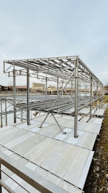 A metal framework structure, likely part of a construction or installation project, is situated near a body of water. The structure comprises numerous horizontal and vertical metal beams, set against an urban backdrop with buildings visible in the distance.