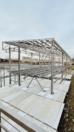 A metal framework structure, likely part of a construction or installation project, is situated near a body of water. The structure comprises numerous horizontal and vertical metal beams, set against an urban backdrop with buildings visible in the distance.