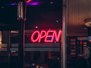 A neon sign with the word 'OPEN' glows in bright red against a dark background. The sign is positioned on a window or glass door, with the interior of a dimly lit establishment visible in the background.
