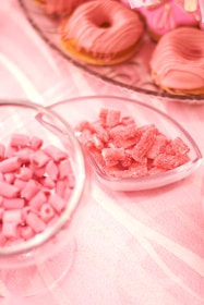 An assortment of pink-themed sweets includes frosted donuts, gummy candies, and small candy rolls arranged on a light pink tablecloth.