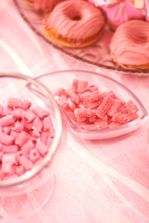 An assortment of pink-themed sweets includes frosted donuts, gummy candies, and small candy rolls arranged on a light pink tablecloth.