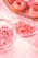 An assortment of pink-themed sweets includes frosted donuts, gummy candies, and small candy rolls arranged on a light pink tablecloth.