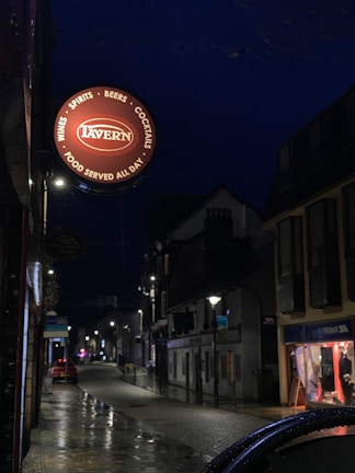 A dimly lit street scene at night features a prominent circular sign for a tavern. The sign includes words like 'Wines,' 'Spirits,' 'Beers,' 'Cocktails,' and 'Food Served All Day.' The street appears wet, suggesting recent rain, and is lined with illuminated street lights and small shops.