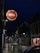 A dimly lit street scene at night features a prominent circular sign for a tavern. The sign includes words like 'Wines,' 'Spirits,' 'Beers,' 'Cocktails,' and 'Food Served All Day.' The street appears wet, suggesting recent rain, and is lined with illuminated street lights and small shops.