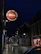 A dimly lit street scene at night features a prominent circular sign for a tavern. The sign includes words like 'Wines,' 'Spirits,' 'Beers,' 'Cocktails,' and 'Food Served All Day.' The street appears wet, suggesting recent rain, and is lined with illuminated street lights and small shops.
