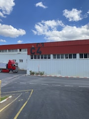 A red and white industrial building with the letters 'C4' prominently painted on the upper section. A red truck is parked beside the building on a paved area, and there is a small speed limit sign indicating 20 km/h nearby. The sky is blue with scattered fluffy clouds.