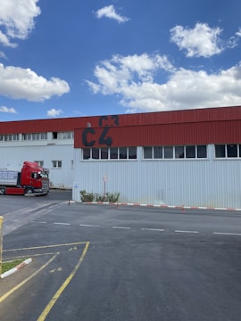A red and white industrial building with the letters 'C4' prominently painted on the upper section. A red truck is parked beside the building on a paved area, and there is a small speed limit sign indicating 20 km/h nearby. The sky is blue with scattered fluffy clouds.