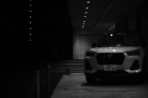 A black and white photograph of a modern SUV parked in front of a building with a garage door. The scene is dimly lit, with overhead lights creating a row of small, bright spots leading into the background. The image has a stark contrast between light and shadow, emphasizing the sleek lines of the vehicle.