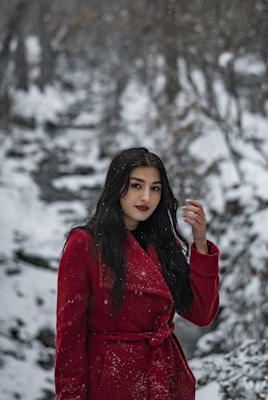 A woman with long dark hair stands in a snowy winter landscape. She is wearing a bright red coat with snowflakes scattered on it. The background features a blurred, snow-covered forest path lined with bare trees.