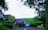 A quaint countryside house with a stone facade and a partial pink extension is surrounded by lush greenery, large trees, and a spacious grassy field. The road in front of the house appears to be paved, with well-maintained bushes bordering the property. The sky overhead is overcast.