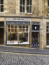 A charming café storefront with large windows displaying a cozy interior and various items. A sign on the facade reads 'The Milkman'. The building is made of stone with a traditional design. The street outside is cobblestone, and there is a gentle slope leading up to the cafe's entrance.